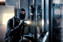 Burglary. Skilful Professional Masked Burglar Opening A Window And Holding A Torch And Breaking Into The House