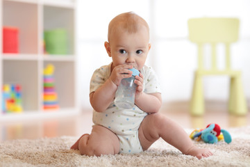 Pretty baby boy drinking water from bottle. Kid sitting on carpet in nursery at home. Smiling child is 7 months old.