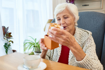 Sticker - Waist-up portrait of pretty senior woman wearing knitted cardigan sitting at cafe table and opening small gift box while celebrating birthday