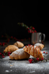 several whole croissants with crumbs, powdered sugar, caramel, a glass with berries and red berries on a dark background