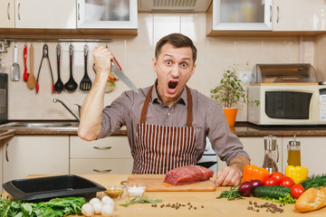 Wall Mural - Angry upset caucasian young man in apron sitting at table with vegetables, cooking at home preparing meat stake from pork, beef or lamb, in light kitchen with wooden surface, full of fancy kitchenware