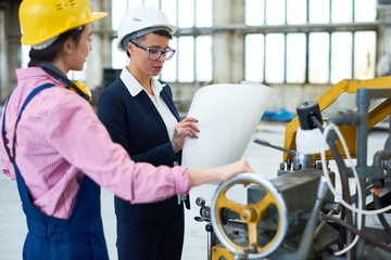 Portrait of two young women wearing hardhats  standing in workshop and holding construction plans, surveying work process at modern plant, copy space