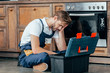 © LIGHTFIELD STUDIOS - tired young foreman sitting with toolbox near broken oven