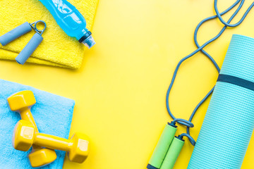 Fitness background. Equipment for gym and home. Jump rope, dumbbells, expander, mat, water on pastel yellow background top view copy space