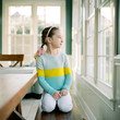 © Jakob/Stocksy - Cute young girl in a colorful sweater sitting on a bench by kitchen table looking outside