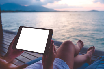 Mockup image of a woman using black tablet pc with blank white desktop screen while sitting by the sea with blue sky background