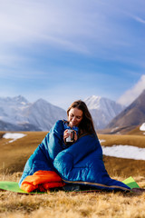 a cute young girl sits in a blue sleeping bag against the background of the Caucasus mountains, drinks hot tea, laughs and enjoys nature