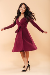 Wall Mural - Full size portrait of charming asian female in pretty maroon dress dancing and looking on camera being isolated over beige background