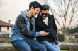 © theartofphoto - Two trendy casual young men sitting together on a bench outdoors reading an sms or text message, or looking at photos on a mobile phone