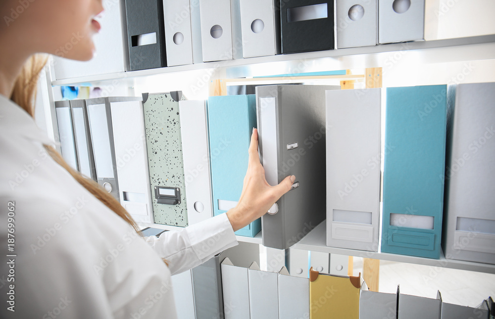 Young woman taking folder with documents from shelf in archive