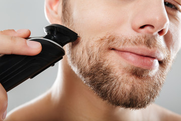 Canvas Print - Close up portrait of handsome man being contented and happy while shaving his skin with electric shaver in morning against grey wall