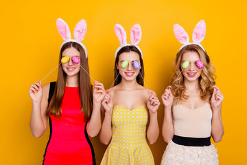 Wall Mural - Happy Easter! Three pretty, trendy, comic, funny girls wearing bunny ears on heads in dresses closed eyes with easter eggs, standing over yellow background