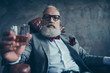© deagreez - Attractive, old investor in spectacles, hold glass with brandy, in tuxedo with red bowtie and pocket square, sit in leather chair over gray background, looking at the camera, shares, stock, money