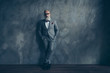 © deagreez - Full length portrait of stunning perfect brutal harsh old man in suit with bow-tie holding hands in pockets, leg by foot standing on wooden floor over gray wall