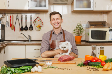 Wall Mural - Young man in apron sitting with furry cat at table with vegetables, cooking at home preparing meat whitestake from pork, beef or lamb, in light kitchen with wooden surface, full of fancy kitchenware.