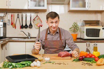 Wall Mural - Attractive caucasian young man in apron sitting at table with vegetables, cooking at home preparing meat stake from pork, beef or lamb, in light kitchen with wooden surface, full of fancy kitchenware.