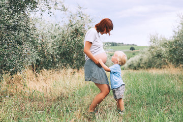 Wall Mural - Child boy kissing belly of pregnant her mother on nature background.