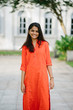 © Danon - Portrait of young indian woman wearing bright orange ethnic wear. She is standing outdoors with a white background and is smiling at the camera.