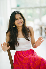 Wall Mural - Portrait of young and attractive Indian woman in a white top sitting in a wood chair and smiling radiantly 