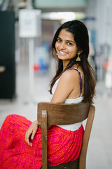 Portrait of young and attractive Indian woman in a white top sitting in a wood chair and smiling radiantly 