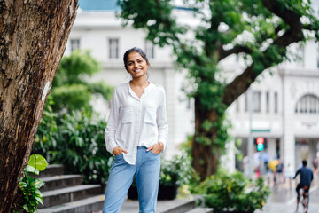 Wall Mural - Candid and authentic portrait of a smiling, attractive and young Indian woman standing in the sun next to a tree in a city with traffic in the background.