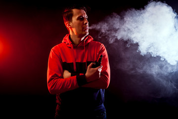 Young stylish male smoker in a sweatshirt  blows out big cloud of smoke from vape on a dark isolated background