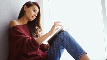 Poster - Side view of Pleased brunette woman in sweater sitting near the window and using tablet computer at home
