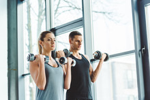 Focused Young Man And Woman Exercising With Dumbbells In Gym