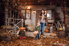 Cute Little Baby Boy And Mother Dressed In A Sweater, Jeans Playing Near House With Plush Toy Teddy Bear In Autumn Time. Woman And Son On Courtyard, Lit By Flashlights, With Dry Fall Leaves, Pumpkins.