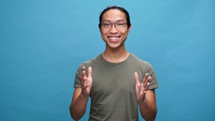 Poster - Surprised happy asian man in t-shirt and eyeglasses rejoice and looking at the camera over blue background