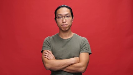 Poster - Displeased asian man in t-shirt and eyeglasses with crossed arms waving his head and looking at the camera over red background