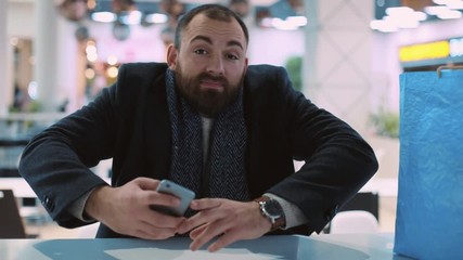 handsome young man use phone sitting look at camera smiling shrugs in mall feel happy close up attractive waiting shopping look around portrait new year holiday smartphone internet slow motion