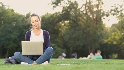 Poster - Pleased brunette woman sitting on grass and using laptop computer in park