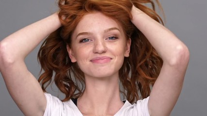 Poster - Close up view of funny ginger woman in t-shirt playing with her hair and looking at the camera over gray background