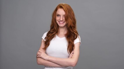 Wall Mural - Smiling mystery ginger woman in t-shirt holding crossed arms and looking at the camera over gray background
