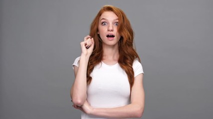 Wall Mural - Serious pensive ginger woman in t-shirt holding her chin and having idea over gray background
