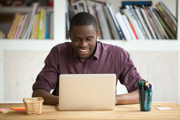 Wall Mural - Smiling african-american man using laptop sitting at home office desk, happy excited black e business owner looking at computer screen pleased by reading good news, chatting in social network