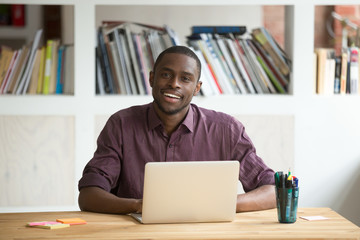 Wall Mural - Portrait of friendly attractive african-american businessman entrepreneur sitting at desk with laptop looking at camera, smiling black man business professional posing at workplace with computer