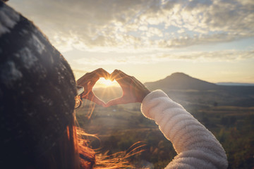 young woman traveler making heart shape symbol at sunrise