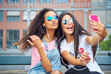 Smiling young girlfriends sitting outside taking selfies together