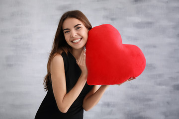 Romantic young woman with heart-shaped pillow on light background