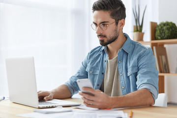 Photo of serious focused man with beard and mustache uses modern digital smart phone device, chats in social networks, keyboards on laptop computer, reads news on websites, makes payment online