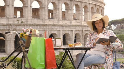 Young woman tourist reads a book sitting at the table outside a bar restaurant in front of the Colosseum in Rome with coffee, juice and cornetto. Elegant beautiful dress with large hat and colorful