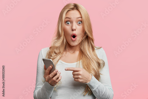 Fotografía Blonde young female with pleasant appearance looks with terrified expression in smart phone, reads shocking news on webpage, isolated over pink background