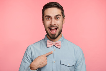 Wall Mural - Puzzled young male model indicates at himself with indignant expression, demonstrates new bowtie, isolated over pink background. Handsome man looks in bewilderment at camera, gestures indoor