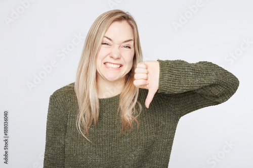 Fotomural European blonde stylish woman keeps thumb down, disapproves something, frowns face in discontent