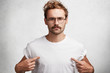 © wayhome.studio - Photo of serious confident fashionable male with trendy hairdo, wears round spectacles and white t shirt, indicates at blank copy space of his clothes for your designing content. People and clothing