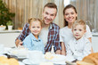 © pressmaster - Young couple and two cute children looking at camera while sitting by dinner table