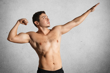 Photo of fit strong young male bodybuilder poses, shows flexed muscles, stretches hands, isolated over grey background. Fitness model with naked torso demonstrates his strength and sporty body