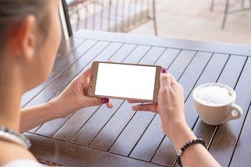 Poster - Woman holding mobile phone in horizontal position with isolated screen for mockup. Coffee cup and wooden desk in background.